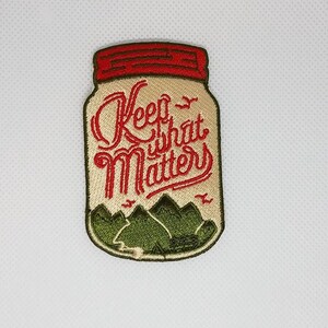 Könnte beinhalten: Gestickter Aufnäher mit einem Einmachglas-Design. Das Glas ist mit grünen Bergen gefüllt und der Text "Keep what matters" ist in roter Kursivschrift geschrieben.