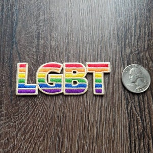 Könnte beinhalten: Ein gestickter Patch in Regenbogenfarben mit den Buchstaben "LGBT" in Weiß. Der Patch ist etwa so groß wie eine 2-Euro-Münze.