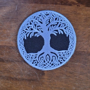 May include: A light blue embroidered patch with a white tree of life design. The tree has a thick trunk and branches with intricate details. The background is black.