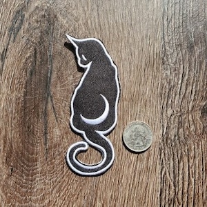 Puede incluir: Parche de hierro bordado negro de un gato con una luna creciente en su cola. El parche está sobre un fondo de veta de madera.