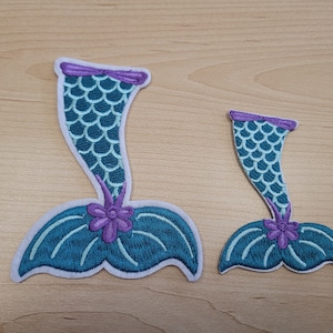 May include: Two embroidered mermaid tail patches. The larger patch is teal blue with purple accents and the smaller patch is teal blue with purple accents.