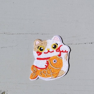 May include: White and brown cat with red and orange details holding a gold fish. The cat is a traditional Japanese Maneki-Neko, or lucky cat, with a raised paw.