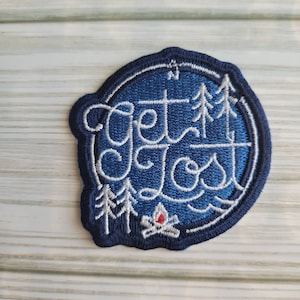 Puede incluir: Un parche bordado azul y blanco con el texto "Get Lost" en una fuente cursiva. El parche presenta una brújula, árboles y una fogata.