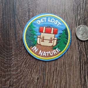Op de afbeelding: Een blauwe en gele geborduurde patch met een rugzak, bomen en de tekst "Get Lost In Nature".