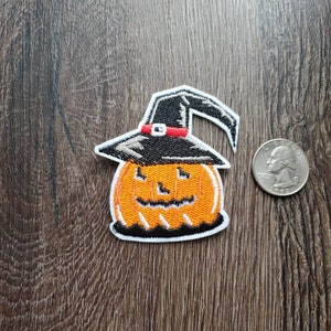 May include: Orange pumpkin with a black witch hat and a smiling face. The pumpkin is embroidered on a white fabric patch.