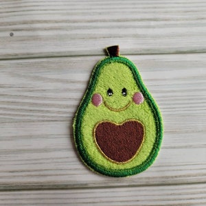 May include: A green felt avocado with a brown heart and pink cheeks. The avocado has a smiling face with black eyes.