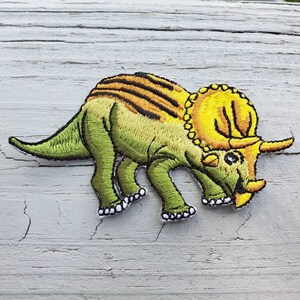 May include: Green and yellow embroidered iron-on patch of a triceratops dinosaur.