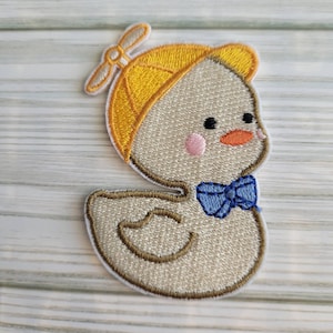May include: Embroidered iron-on patch of a yellow duck wearing a yellow hat with a propeller and a blue bow tie.