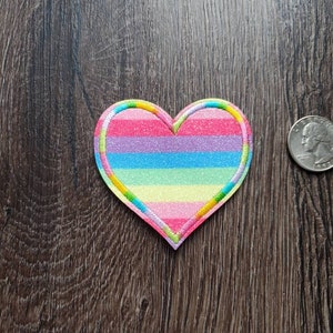 May include: A rainbow striped heart-shaped patch with a glittery finish. The patch is approximately the size of a US quarter.