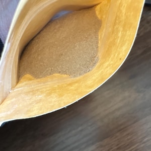 Op de afbeelding: Een open bruin papieren zakje met een lichtbruine, korrelige substantie. Het zakje wordt opengehouden en de inhoud vult het grootste deel van de ruimte. De achtergrond is een donker houten oppervlak.