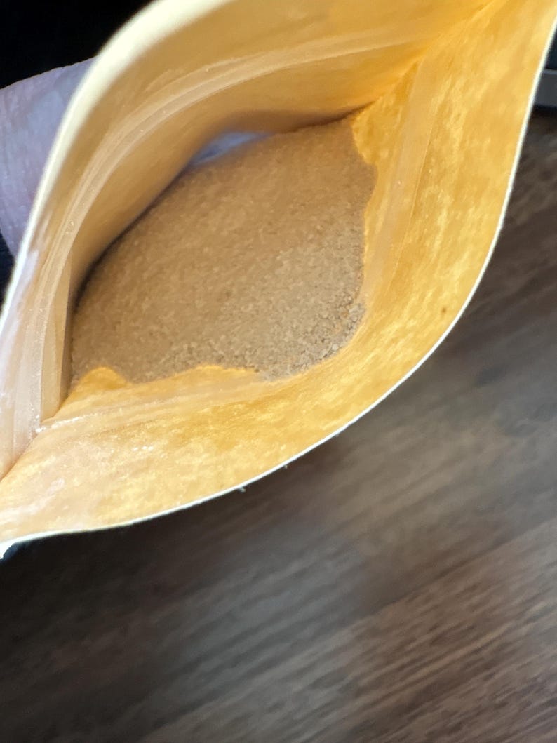 Op de afbeelding: Een open bruin papieren zakje met een lichtbruine, korrelige substantie. Het zakje wordt opengehouden en de inhoud vult het grootste deel van de ruimte. De achtergrond is een donker houten oppervlak.