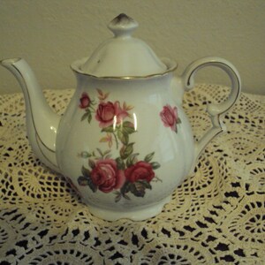 May include: White teapot with a gold trim and a floral design featuring pink roses. The teapot has a lid and a handle.