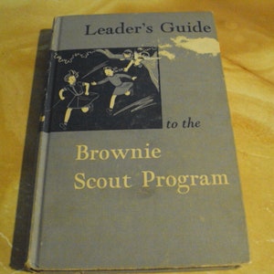 May include: A blue book with the title "Leader's Guide to the Brownie Scout Program". The cover features a black and white illustration of three girls in uniform.