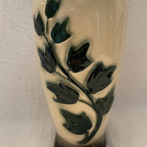May include: A cream-colored ceramic vase featuring a raised, dark green ivy leaf pattern. The vase stands on three small, dark brown feet. A pink measuring tape is visible at the top, indicating a height measurement starting at 8 inches.