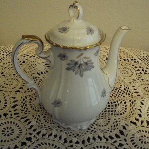 May include: A white porcelain coffee pot with a gold trim and a gray floral design. The pot has a lid and a handle.
