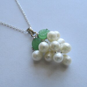 May include: A silver necklace featuring a pendant designed to resemble a bunch of grapes. The pendant includes white pearl beads and green, leaf-shaped accents. The necklace is displayed on a white surface.
