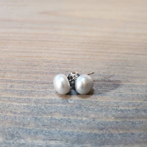 May include: A pair of white pearl stud earrings. The oval-shaped pearls have a glossy finish. The earring posts are silver. The earrings are presented on a light blue and brown wooden surface.
