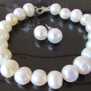 May include: A pearl bracelet and matching stud earrings. The bracelet is made of large, off-white pearls strung together with a silver clasp. The earrings are round, white pearls. The jewelry set is displayed on a dark surface.