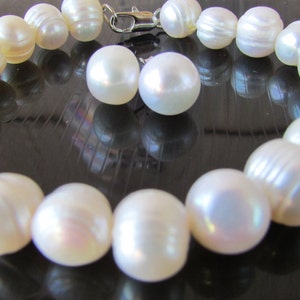 May include: A cream-colored pearl bracelet with a silver clasp and two matching pearl earrings. The bracelet features large, irregularly shaped pearls. The pearls have a lustrous sheen, reflecting light. The jewelry is displayed on a dark, reflective surface.