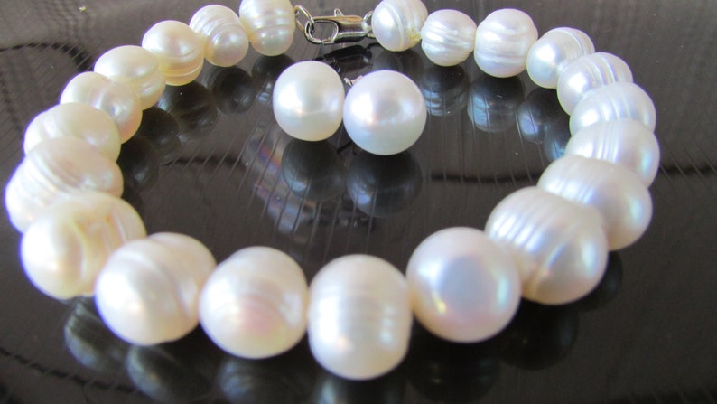 May include: A cream-colored pearl bracelet with a silver clasp and two matching pearl earrings. The bracelet features large, irregularly shaped pearls. The pearls have a lustrous sheen, reflecting light. The jewelry is displayed on a dark, reflective surface.