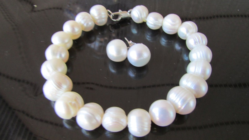 May include: A pearl bracelet and matching stud earrings. The bracelet is made of large, off-white pearls strung together with a silver clasp. The earrings are round, white pearls. The jewelry set is displayed on a dark surface.