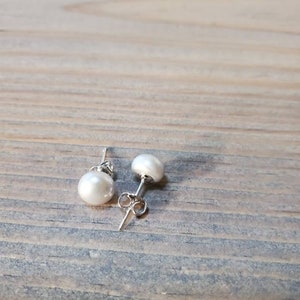 May include: Two white pearl stud earrings with silver posts. The earrings are round and have a lustrous surface. The earrings are displayed on a light brown wooden surface.