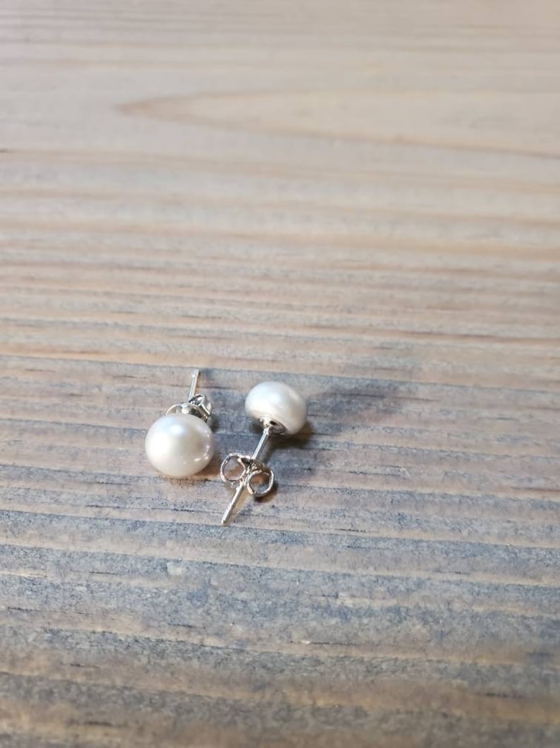 May include: Two white pearl stud earrings with silver posts. The earrings are round and have a lustrous surface. The earrings are displayed on a light brown wooden surface.