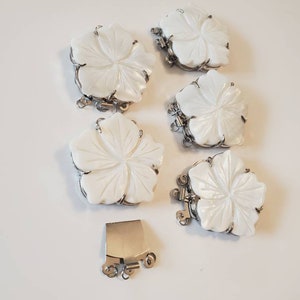 May include: Six white flower-shaped jewelry clasps with silver-colored metal accents. The clasps are carved with petal details and have a shiny, pearlescent finish. One rectangular clasp is also included.