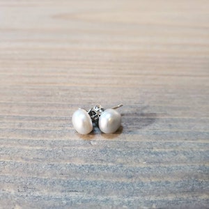 May include: A pair of white pearl stud earrings. The earrings feature oval-shaped pearls and silver posts. The earrings are presented on a wooden surface.