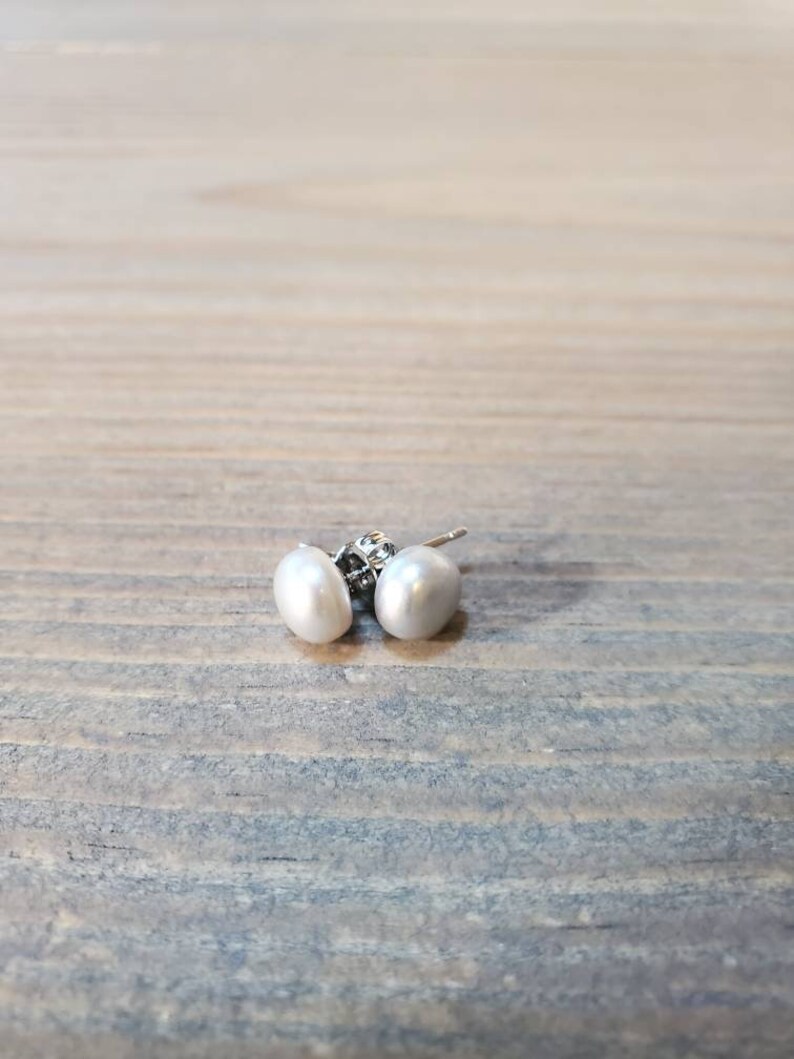 May include: A pair of white pearl stud earrings. The earrings feature oval-shaped pearls and silver posts. The earrings are presented on a wooden surface.
