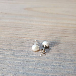 May include: A pair of pearl stud earrings with silver posts. Each earring has a round, white pearl. The earrings are presented on a wooden surface.