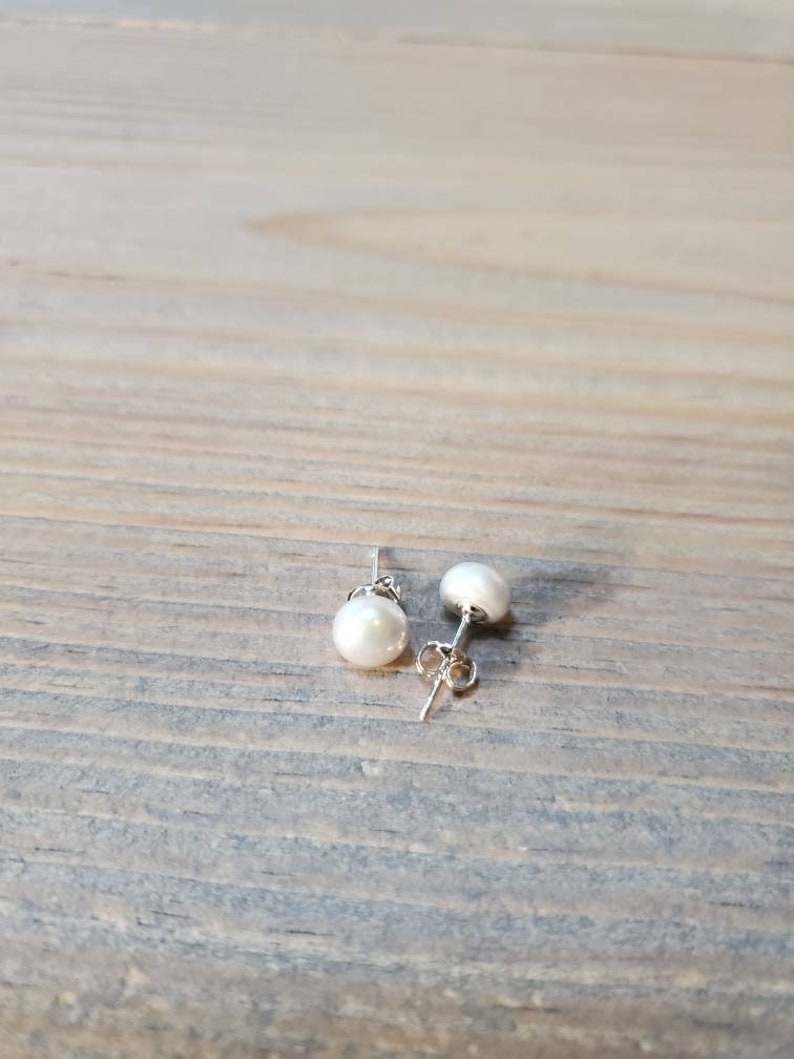 May include: A pair of pearl stud earrings with silver posts. Each earring has a round, white pearl. The earrings are presented on a wooden surface.