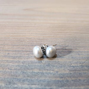 May include: A pair of white pearl stud earrings. The earrings feature oval-shaped pearls and silver posts. The earrings are presented on a wooden surface.