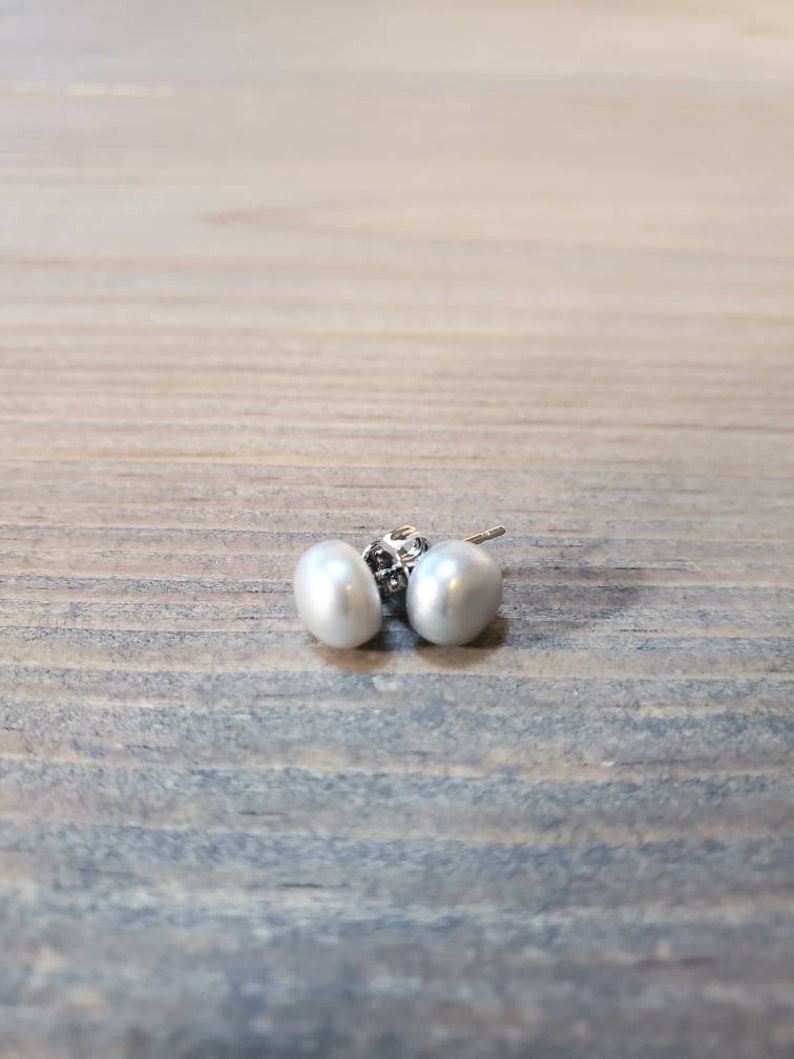 May include: A pair of white pearl stud earrings. The earrings feature oval-shaped pearls and silver posts. The earrings are presented on a wooden surface.