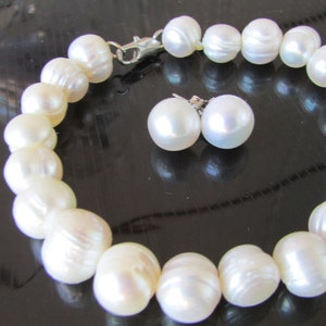 May include: A white pearl bracelet and matching stud earrings. The bracelet is made of large, round pearls strung together with a silver clasp. The earrings are simple, round pearl studs. The jewelry set is displayed on a dark surface.
