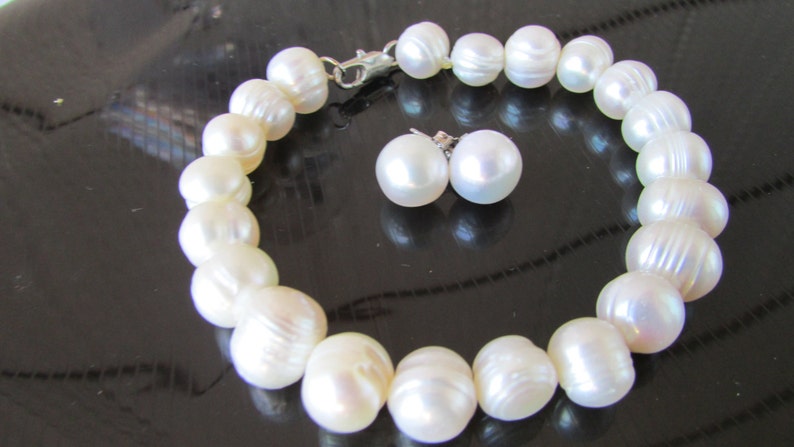 May include: A white pearl bracelet and matching stud earrings. The bracelet is made of large, round pearls strung together with a silver clasp. The earrings are simple, round pearl studs. The jewelry set is displayed on a dark surface.