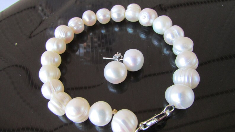 May include: A white pearl bracelet and matching stud earrings. The bracelet is strung with large, irregularly shaped pearls. The earrings feature round, lustrous pearls. The jewelry set is displayed on a reflective black surface.