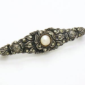 May include: A silver brooch with a pearl center and intricate marcasite detailing. The brooch is shaped like a long, narrow oval.