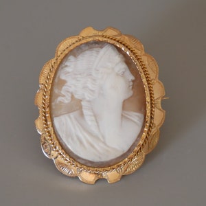 Può includere: Un broche cammeo dorato con una scultura in cammeo bianco di un profilo femminile. Il cammeo è incastonato in una cornice ovale dorata con un bordo a corda.