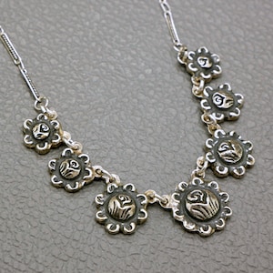 Peut inclure: Un collier en argent avec une chaîne délicate et sept pendentifs en forme de fleur argentés. Chaque pendentif présente un motif en relief complexe.