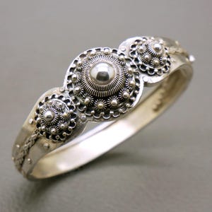 May include: A silver hinged bracelet with three ornate, circular decorative elements. Each element features a central raised sphere surrounded by intricate floral and dot patterns. The bracelet has a polished finish and a secure clasp.