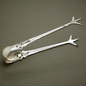 May include: Silver sugar tongs with a decorative floral design and a bird's foot design on the ends.
