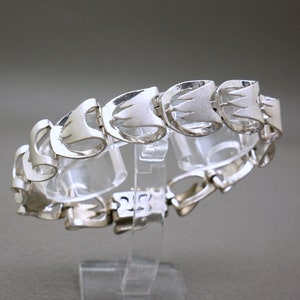 May include: A silver bracelet with a geometric design. The bracelet is made of interlocking links that are shaped like triangles. The bracelet has a clasp closure.