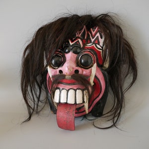 May include: A red, black and white wooden mask with long, black, stringy hair. The mask has a wide, toothy grin and a long, red tongue sticking out. The eyes are large and black, and the nose is small and pink.