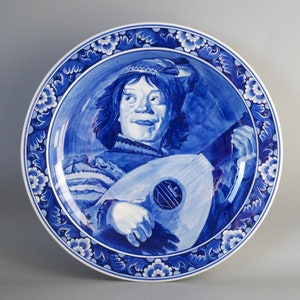 May include: A blue and white ceramic plate with a decorative border featuring floral designs. The center of the plate depicts a person playing a lute, with a blue and white background.