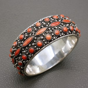 Könnte beinhalten: Ein breiter Silberarmreif mit einem sich wiederholenden Muster aus orangefarbenen Korallen-Cabochons und marquiseförmigen Steinen. Die Metalloberfläche ist mit einer komplexen Granulationsverzierung um jeden Stein versehen.