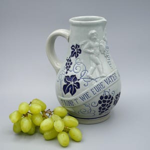 May include: A white ceramic jug with blue and white grapevine designs and a raised relief of a cherub holding grapes. The jug has the text "TRINKT WIE EURE VÄTER" on the side. A bunch of green grapes is in the foreground.