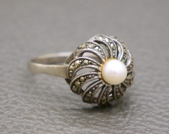 May include: A silver ring featuring a central white pearl surrounded by a decorative, flower-like setting. The setting is embellished with small, sparkling stones, creating a vintage or antique aesthetic. The ring is set against a neutral gray background.