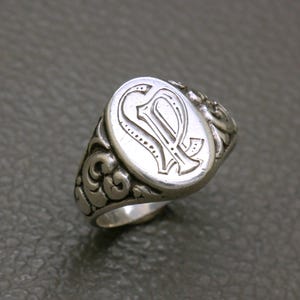 Peut inclure: Une bague chevalière argentée présentant une face ovale avec une lettre P gravée dans une police calligraphiée décorative. L'anneau et les côtés de la bague sont ornés de motifs en relief complexes qui s'étendent depuis le sertissage central.