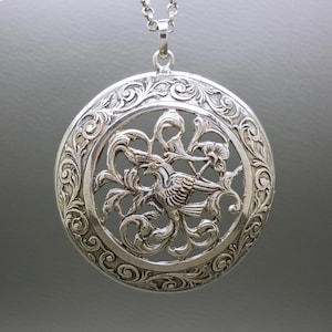 May include: A silver pendant necklace featuring an intricate design of a bird within a floral setting. The pendant is round and has a filigree design.